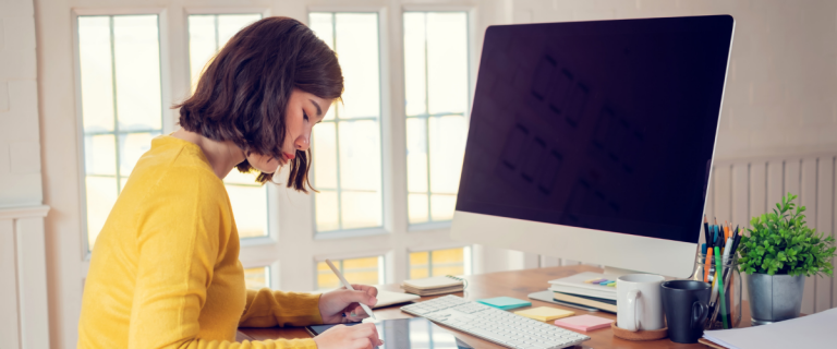 A woman in a yellow sweater works at a desk beside a large desktop screen.