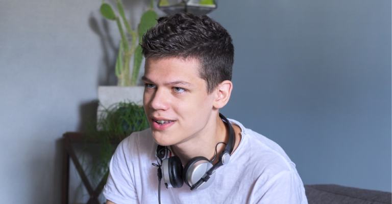 Smiling young man with headphones sits indoors near green plants.